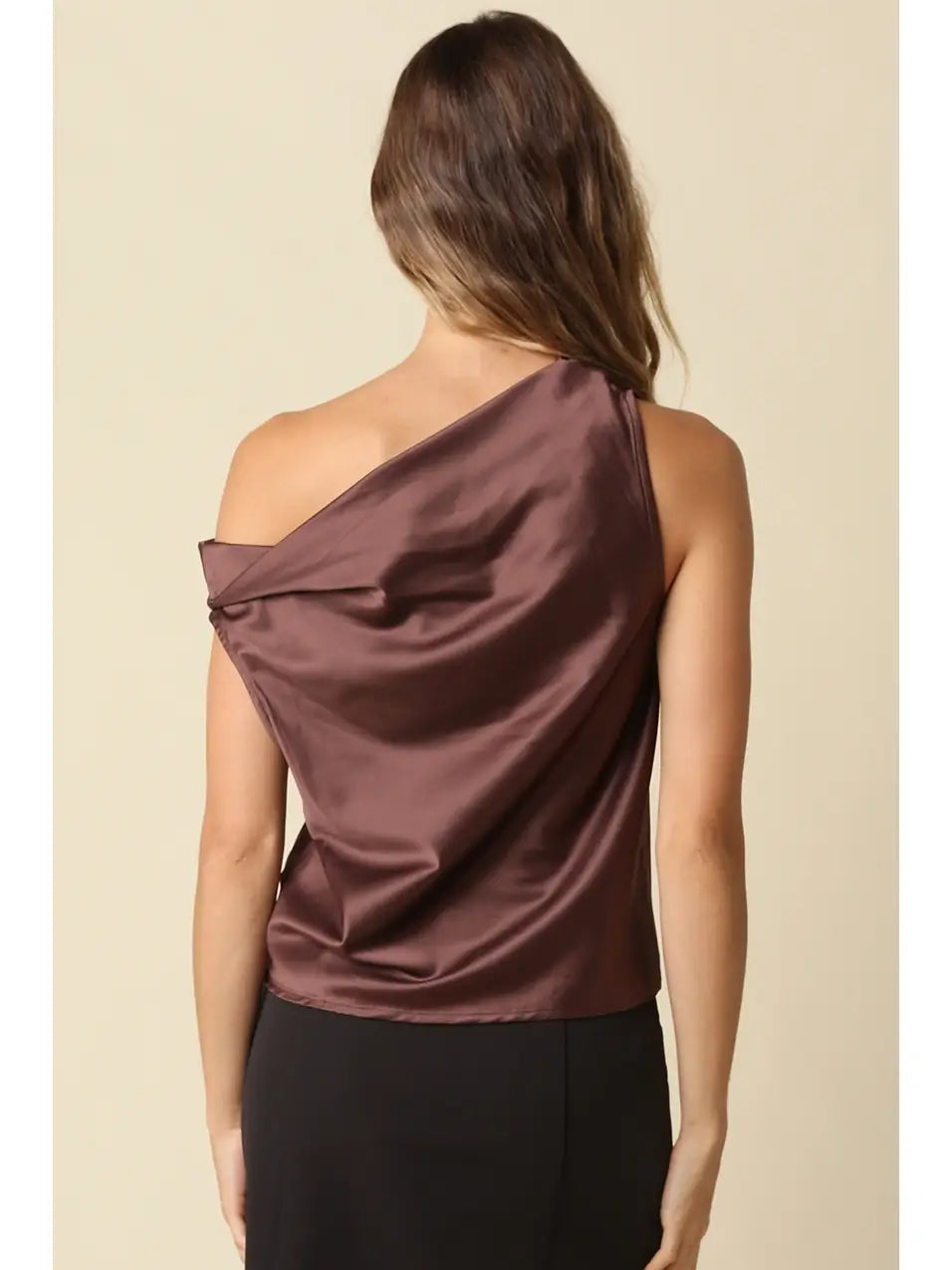 A woman shown from the back wearing a draped one-shoulder satin top in a muted plum-brown color, paired with black bottoms, against a plain beige studio background.