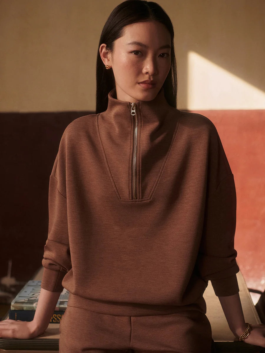 A woman wearing a brown half-zip mock-neck sweatshirt and matching sweatpants, posed in a warm-toned indoor setting with a textured wall behind her.