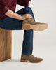 A person seated on a wooden block wearing brown suede cowboy boots with blue jeans and a burgundy shirt, shown from the waist down against a light gray studio background.