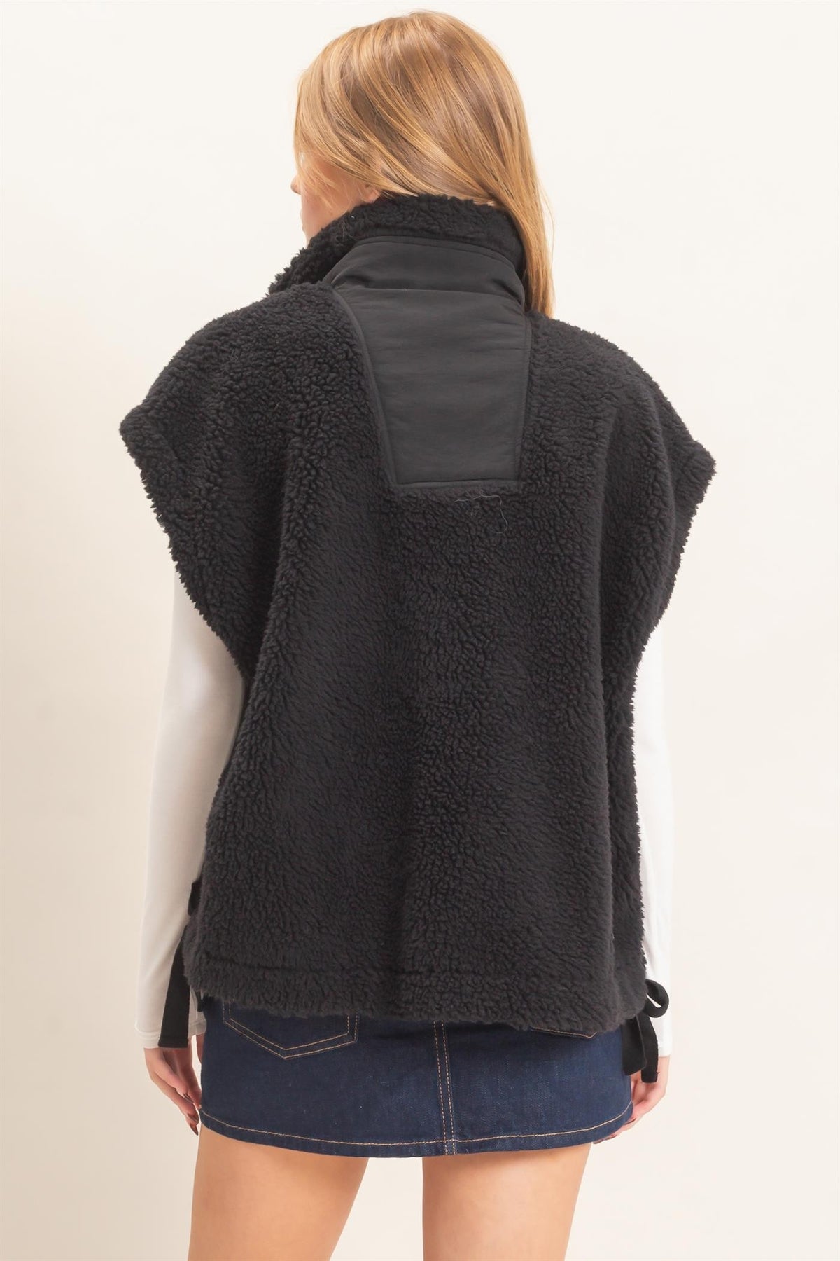 A woman shown from the back wearing a black sherpa fleece vest with a smooth black panel on the upper back, a white long-sleeve shirt, and a dark denim mini skirt against a plain off-white background.