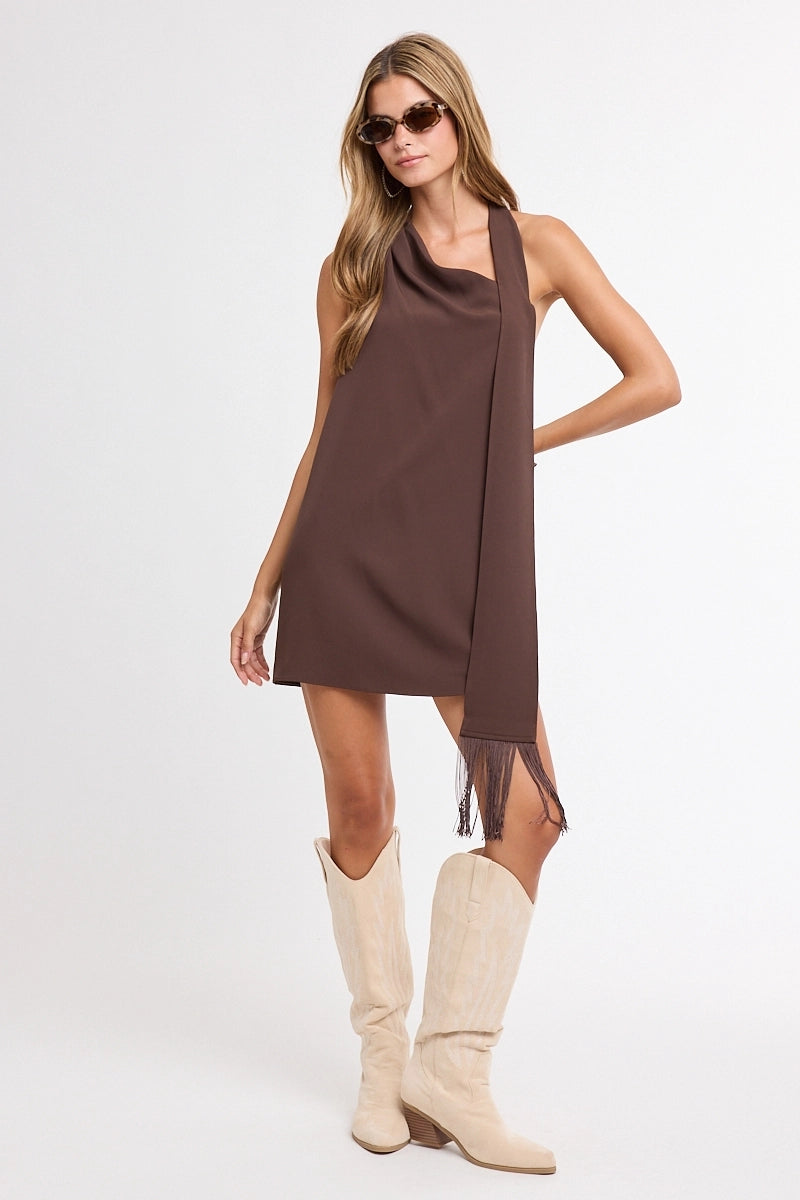A woman modeling a sleeveless, cowl-neck, asymmetrical mini dress in a dark brown color with fringe detailing along the hem, styled with beige cowboy boots and sunglasses against a plain white studio background.