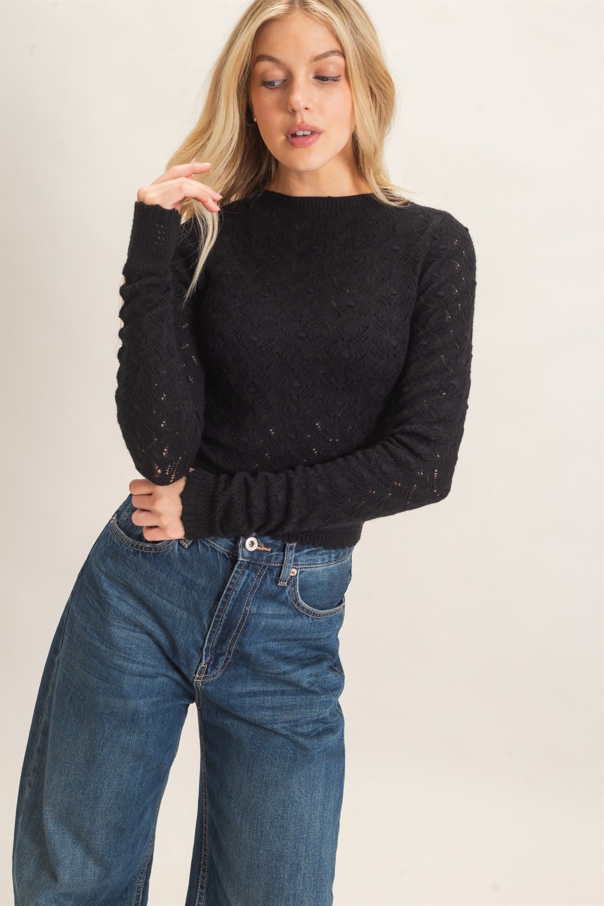 A woman modeling a black long-sleeve knit sweater with textured, slightly sheer detailing on the sleeves and a crew neckline, paired with blue jeans against a plain light background.