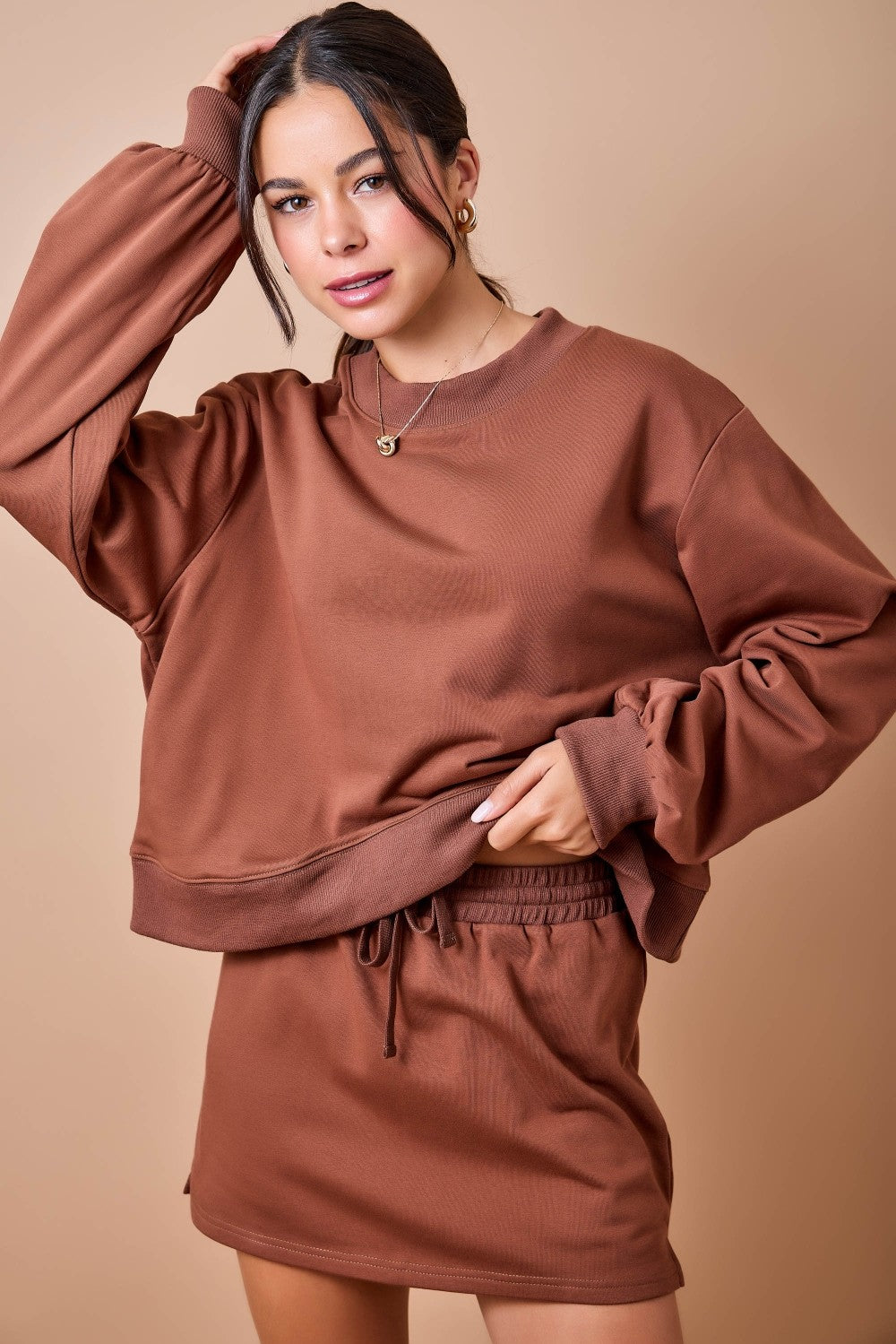 A woman modeling a matching rust-brown lounge set consisting of a long-sleeve sweatshirt and shorts, posed against a solid beige studio background.
