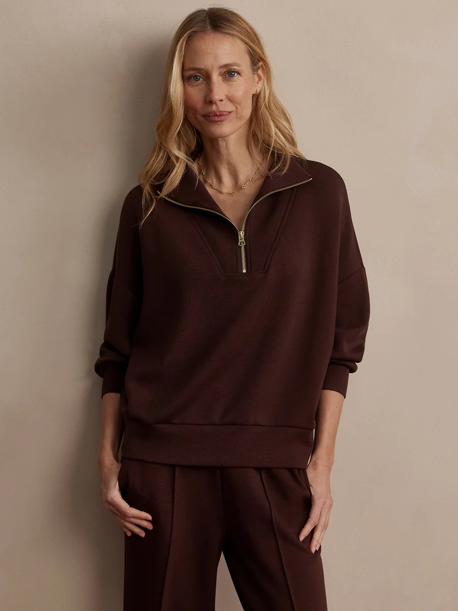 A woman modeling a dark brown half-zip sweatshirt and matching sweatpants against a plain beige studio background.