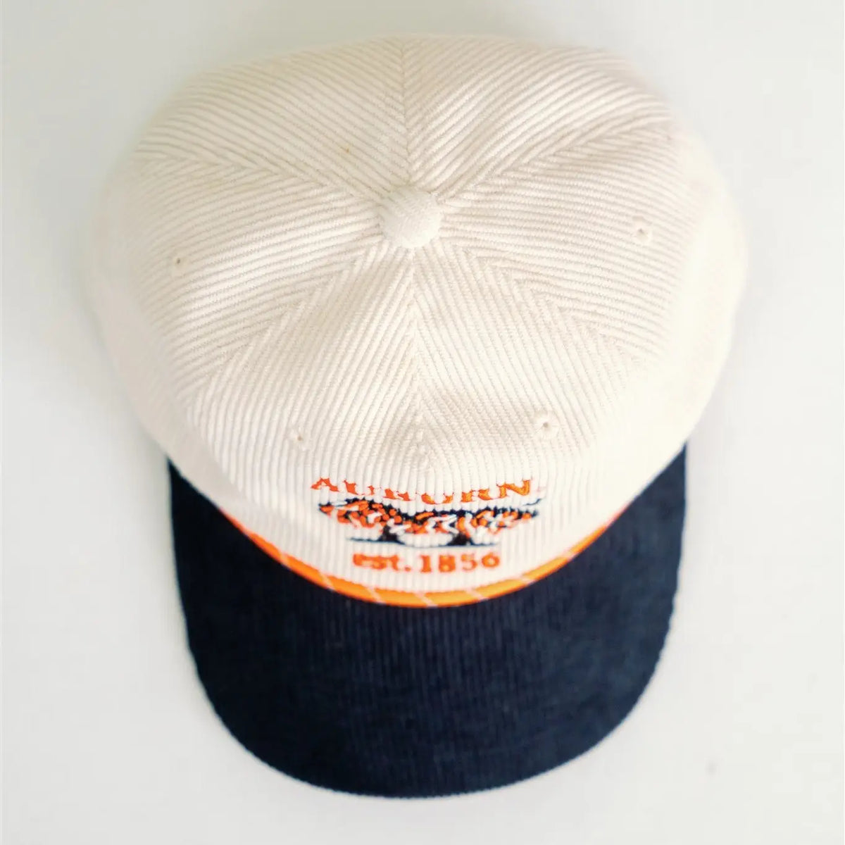 A vintage-style baseball cap with an off-white ribbed crown and a dark navy blue corduroy visor. The front of the cap features orange and white embroidered text reading 'ALVINEN CLOTHING EST.