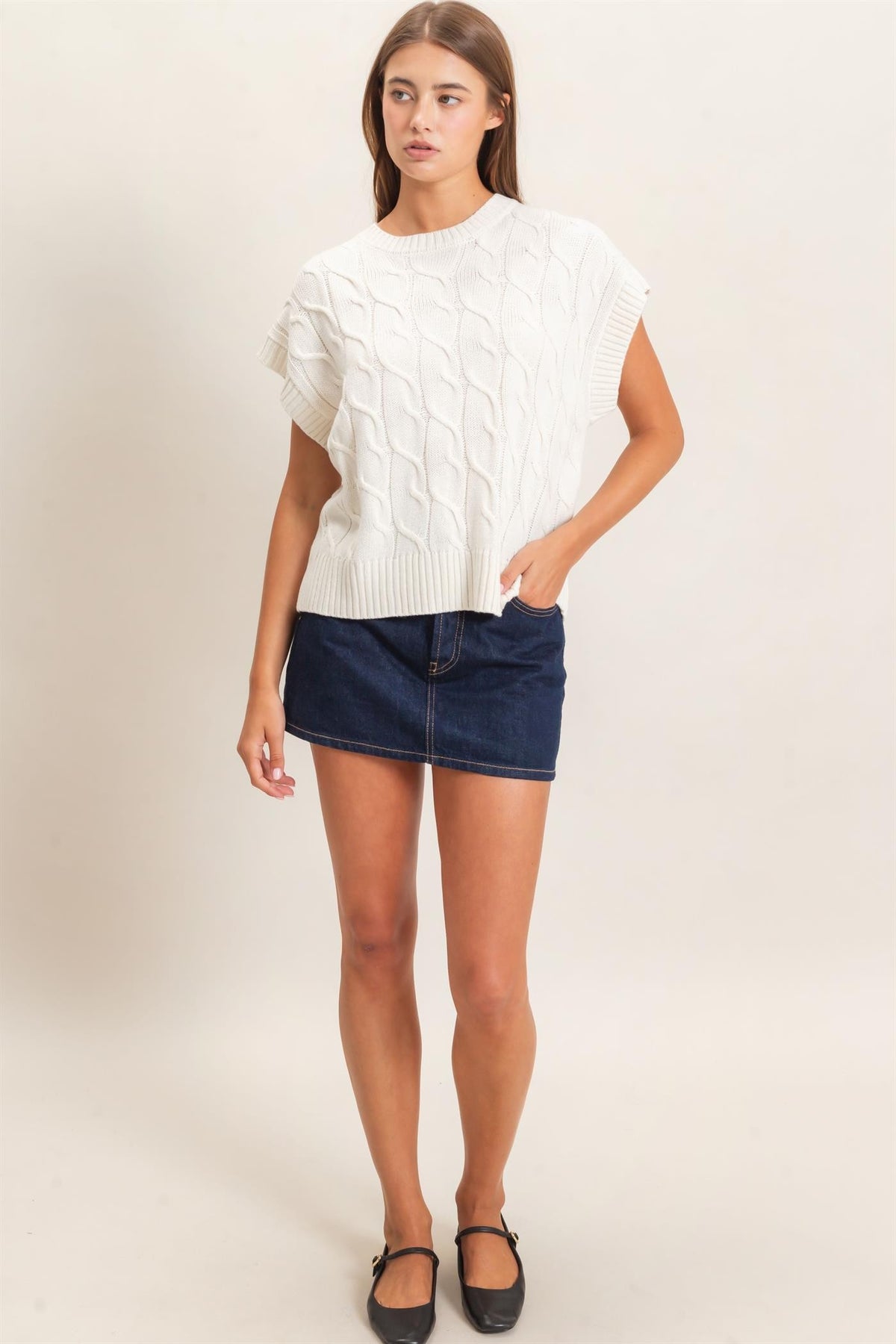A woman modeling a white short-sleeve cable-knit sweater with side cutouts, paired with a dark blue denim mini skirt and black flats, against a plain beige studio background.