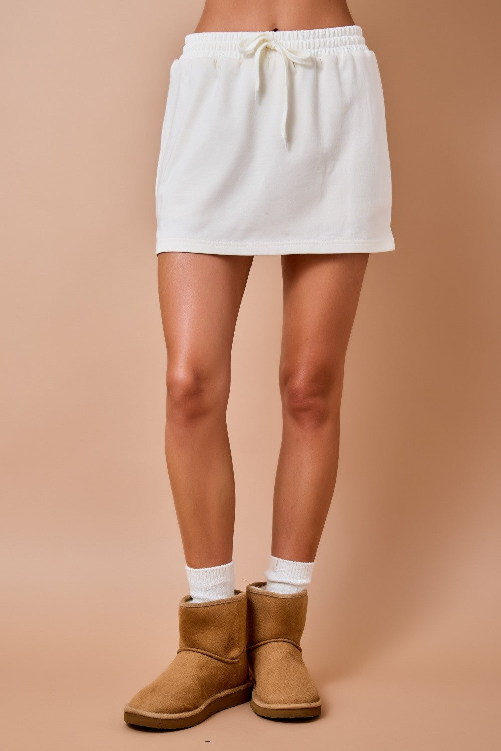A person from the waist down wearing a white drawstring mini skirt, white socks, and tan ankle boots against a solid beige studio background.