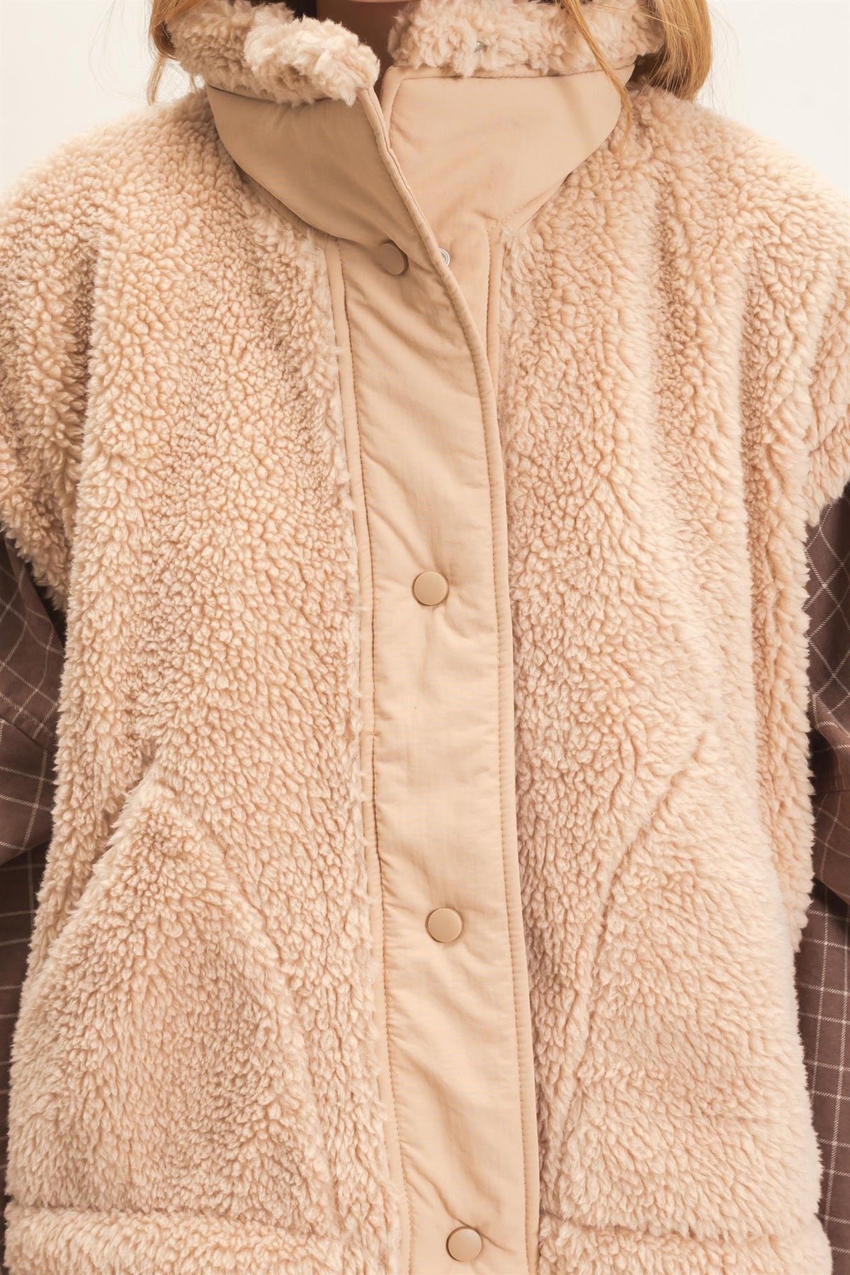 Close-up of a beige sherpa fleece vest with a padded front placket and large buttons, worn over a plaid shirt.