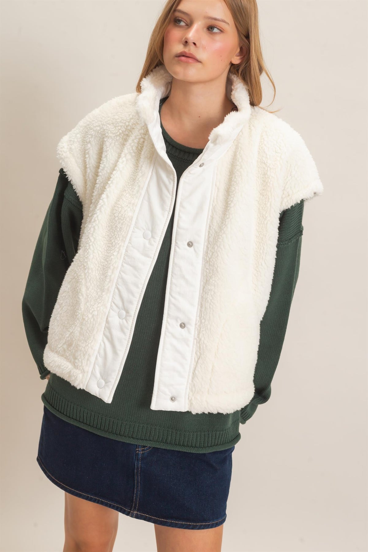 A model wearing a white sherpa-lined button-front vest over a dark green long-sleeve top and a navy denim skirt, photographed against a plain beige studio background.