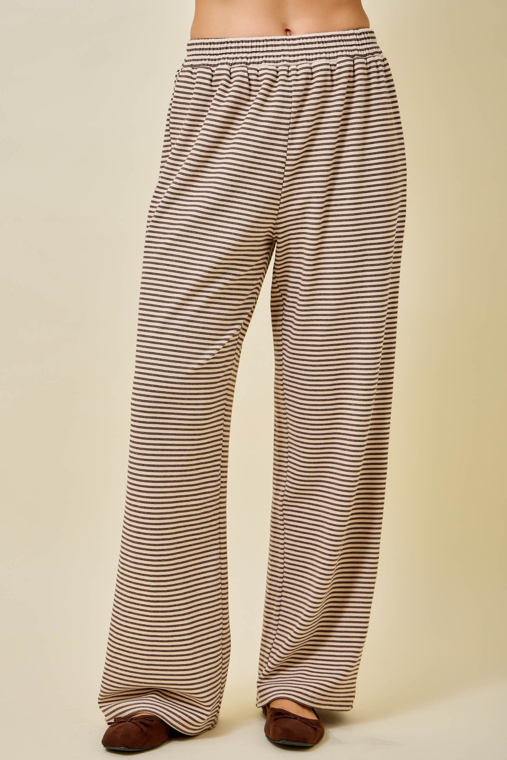 A person wearing loose, wide-leg pants with thin horizontal brown and beige stripes, shown from the waist down against a plain beige studio background.