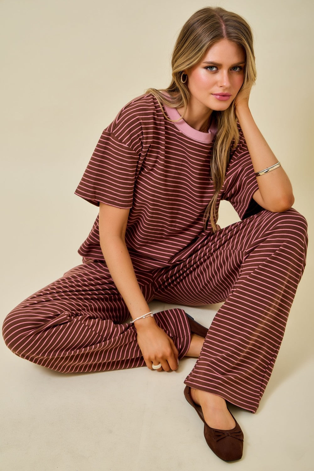 A woman sits cross-legged on a plain beige studio background wearing a matching two-piece lounge set with thin reddish-brown and white stripes. The set includes a short-sleeve top with a light pink inner collar and wide-leg pants.