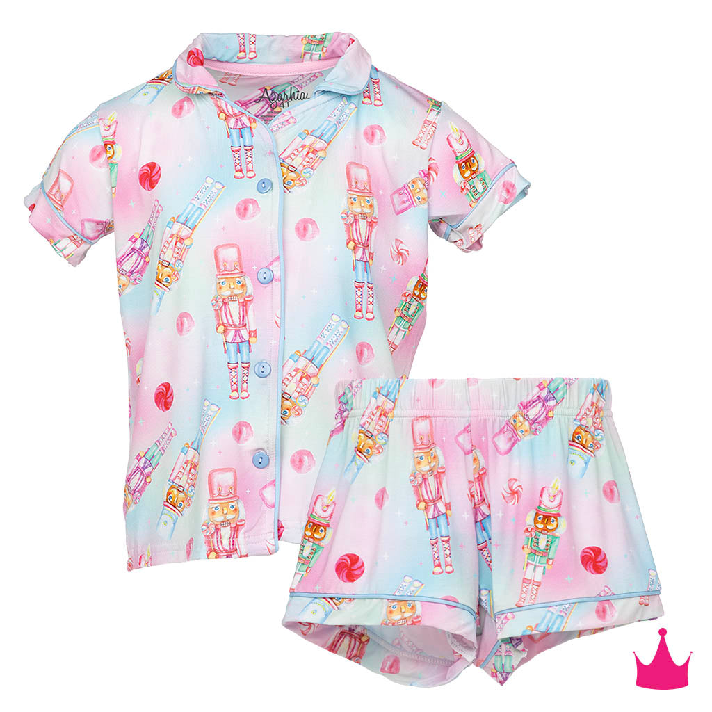 A two-piece children's pajama set featuring a short-sleeve button-front shirt and matching shorts, both printed with a pastel pink and blue candy cane and nutcracker pattern on a white background.