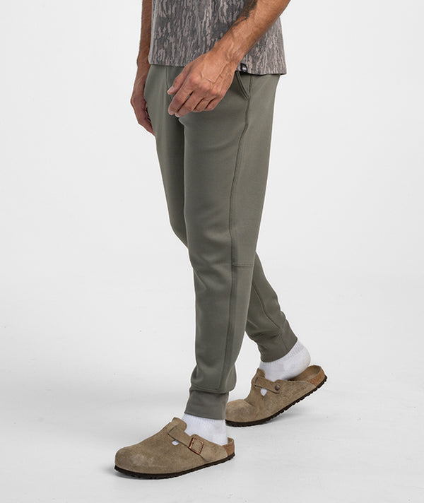A person wearing olive green jogger-style pants, white socks, and tan clogs, shown from the waist down against a clean light gray studio background.