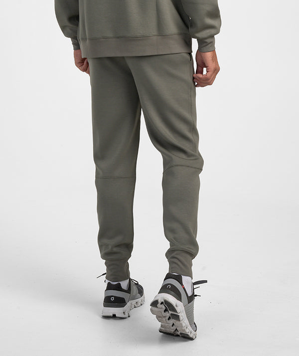 A person shown from the back wearing olive green sweatpants and a matching sweatshirt, paired with gray and white sneakers, against a light gray studio background.