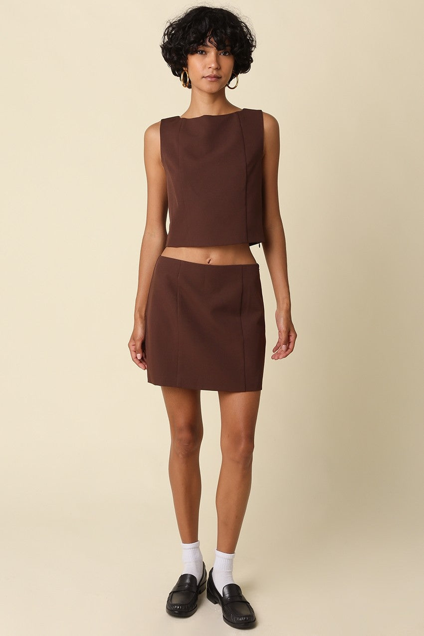 A model stands against a solid beige studio background wearing a matching dark brown sleeveless crop top and high-waisted mini skirt set, styled with white socks and black loafers.