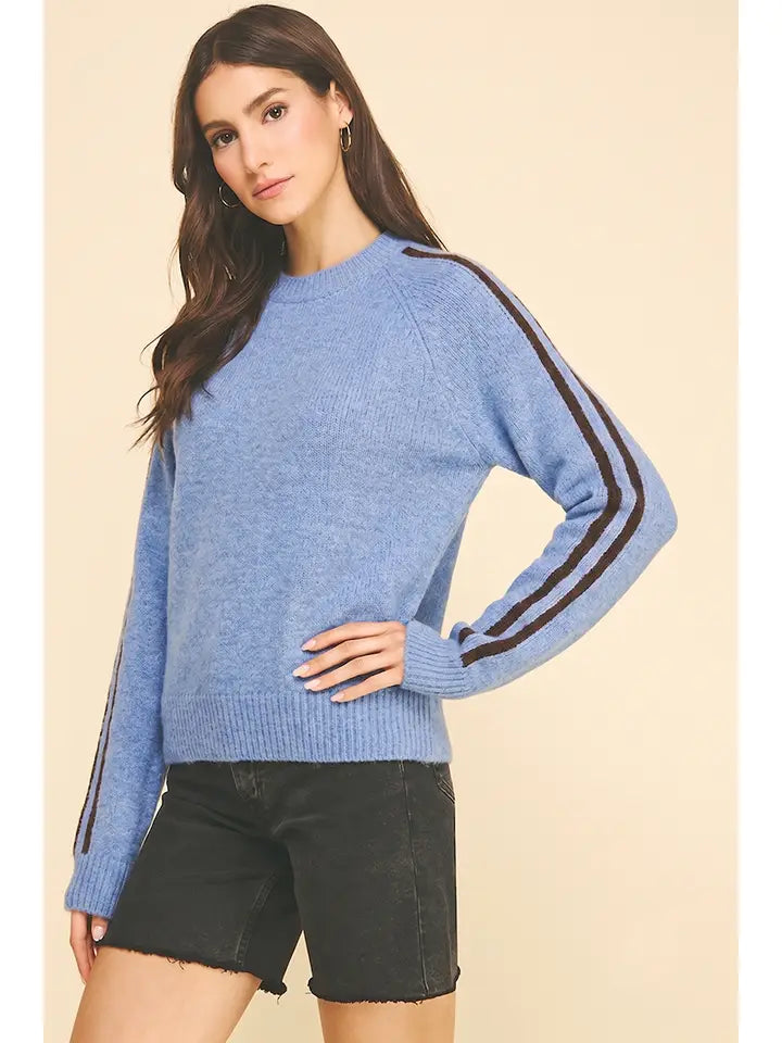 A woman modeling a light blue crewneck sweater with dark brown double stripes down the sleeves, paired with black denim shorts, against a solid beige background.