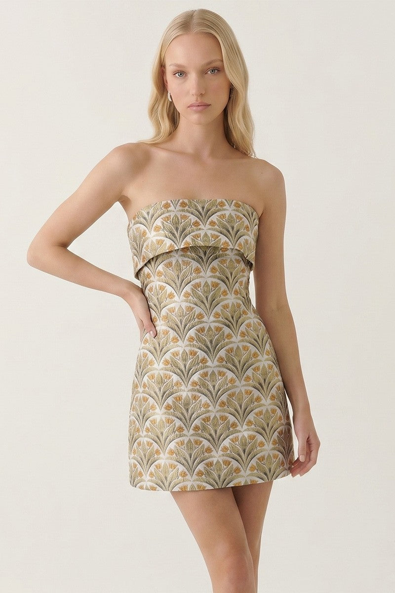 A blonde model wearing a strapless mini dress with a beige, gold, and white scalloped fan pattern against a plain off-white background.