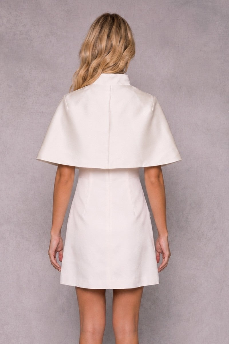 A blonde model wearing a white satin mini dress with a square neckline, thin shoulder straps, and two front flap pockets, and matching cape posed against a textured gray studio background.