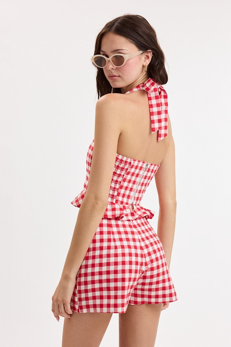 A woman modeling a red and white gingham halter romper from the back, featuring a tie-neck detail and ruffled waist, set against a plain white studio background.