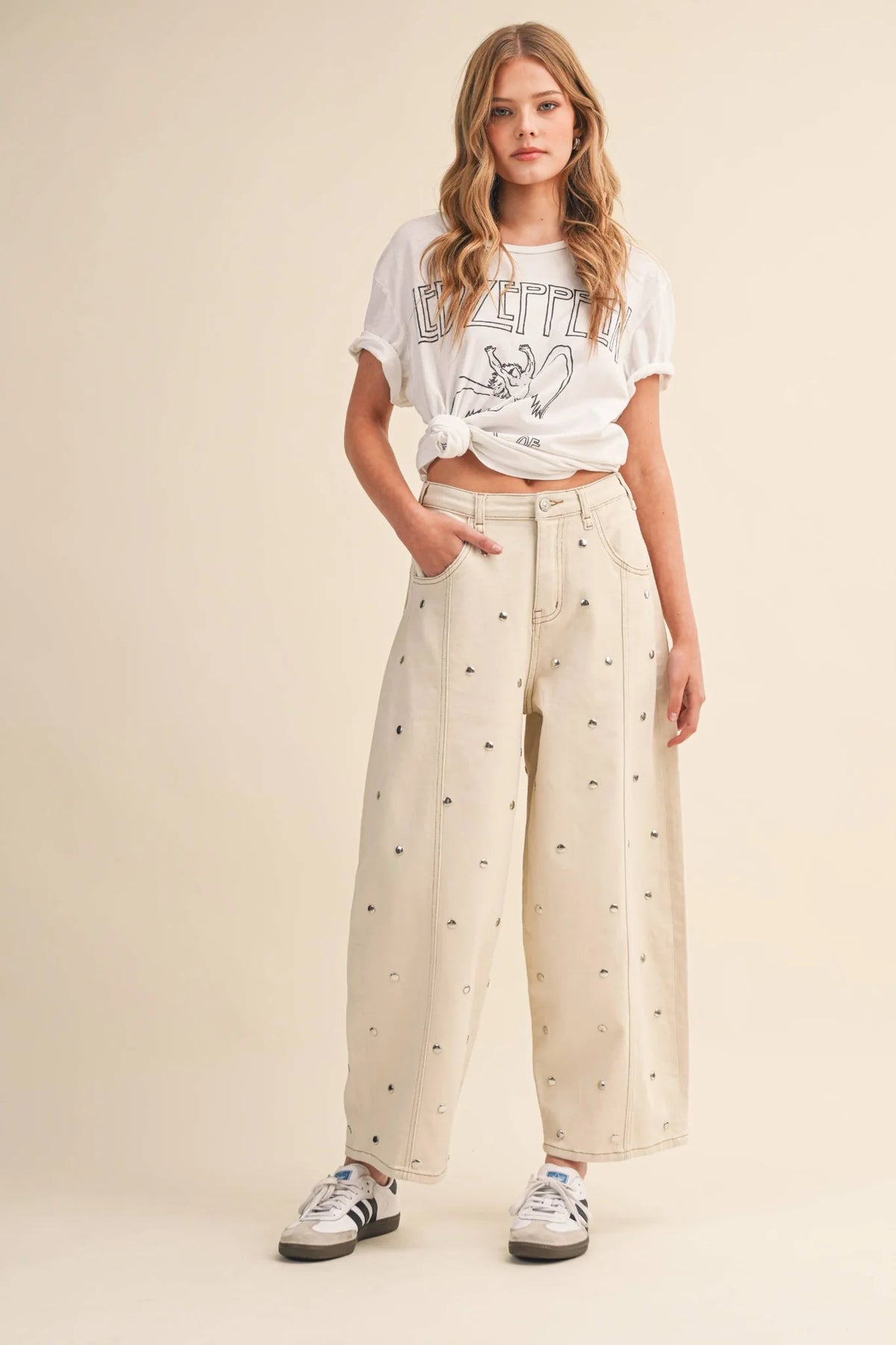 A woman modeling a white graphic t-shirt tied at the waist with cream wide-leg pants featuring a small black pattern, and white sneakers, set against a solid beige studio background.