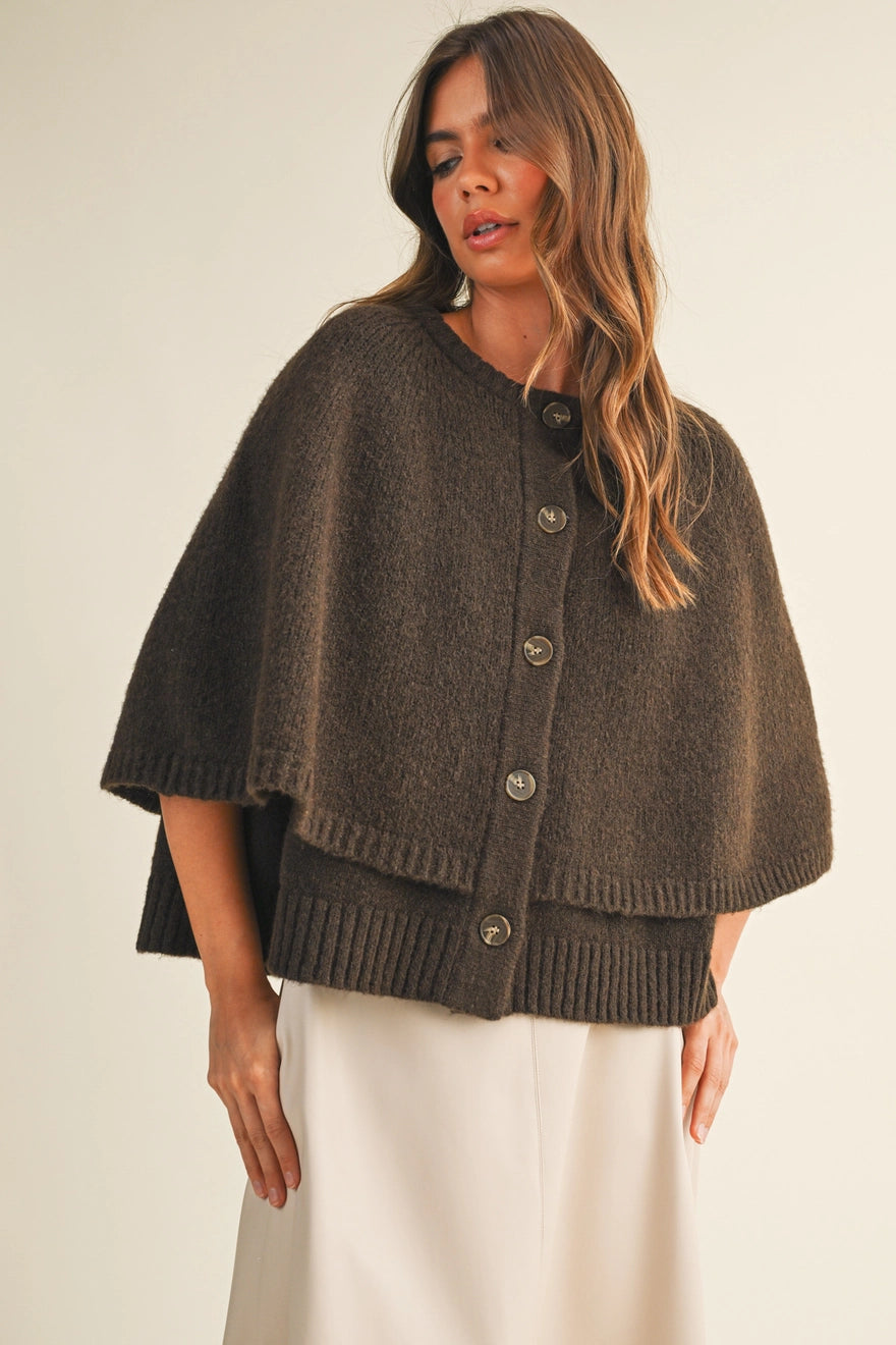 A woman modeling a dark brown textured knit cape cardigan with button-front closure and a cream-colored skirt, set against a plain beige studio background.
