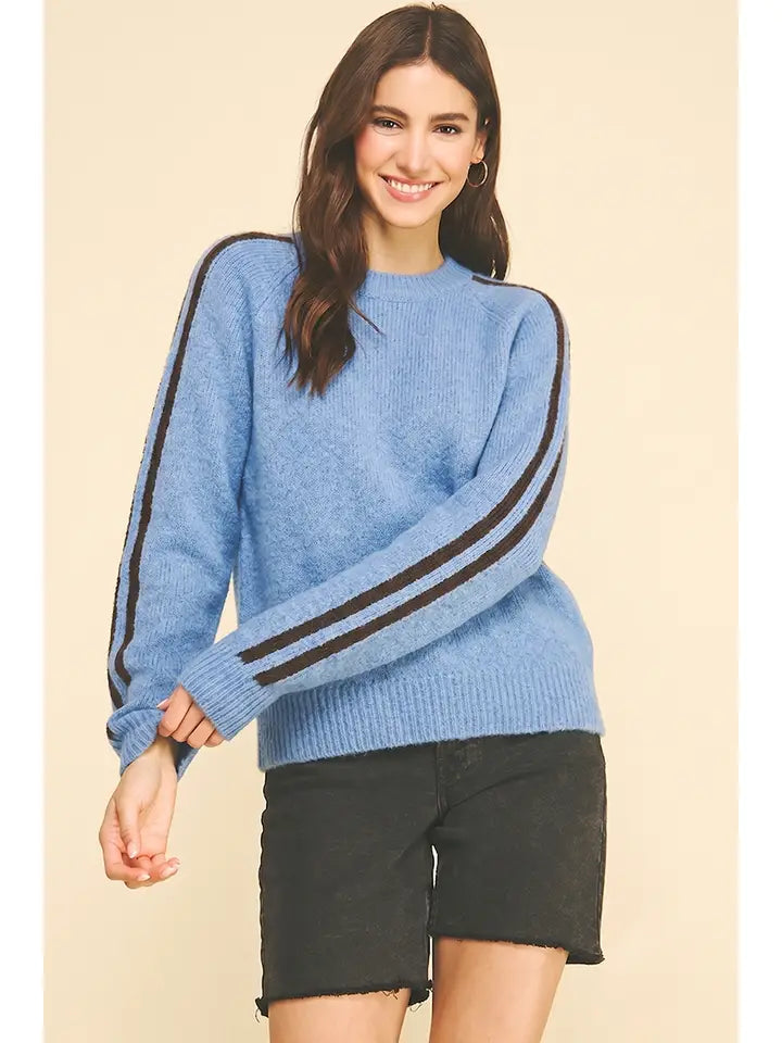 A woman modeling a light blue knit sweater with two dark brown stripes running down the sleeves, paired with black denim shorts, against a solid beige background.