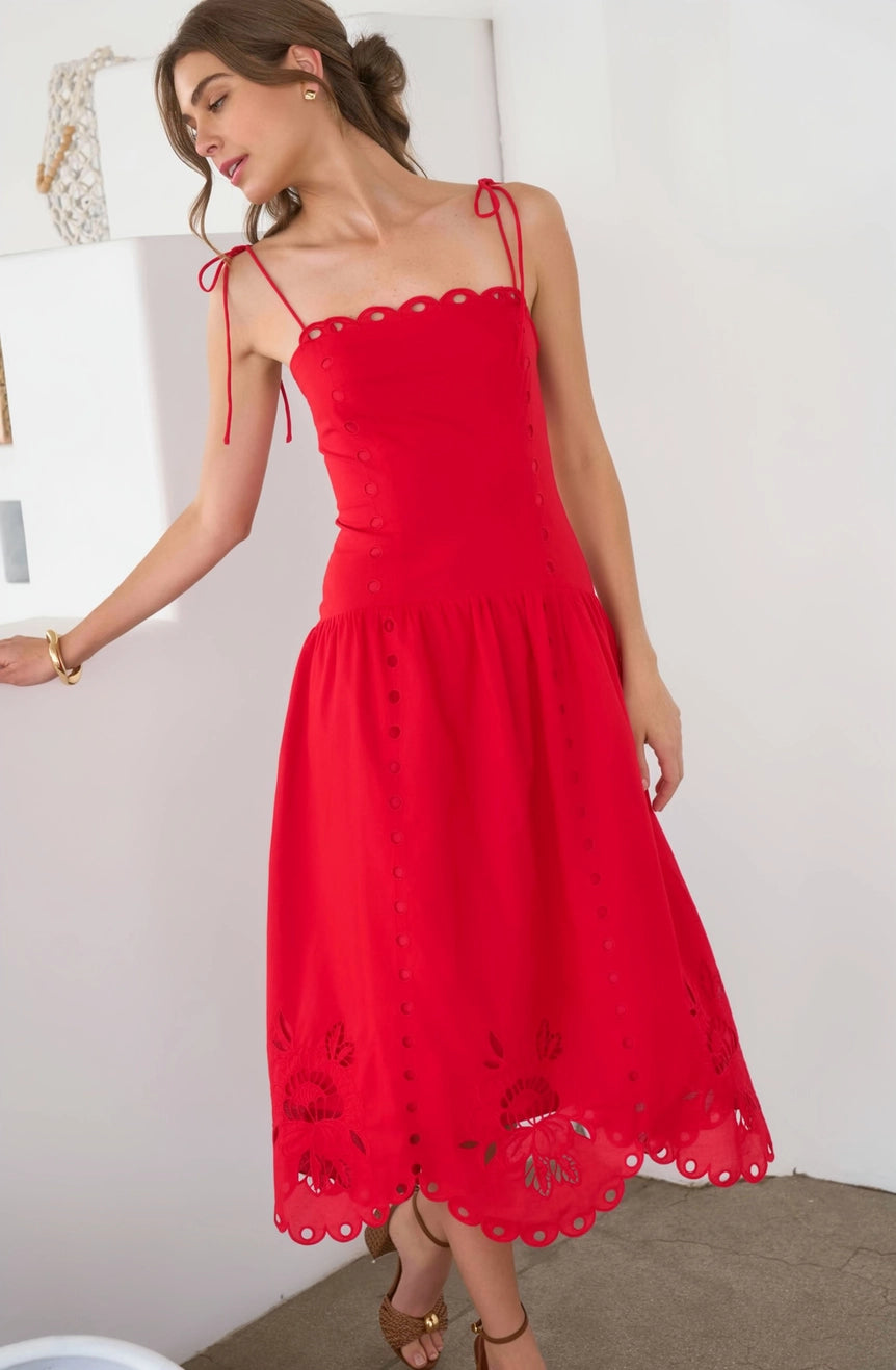 A woman wearing a bright red midi dress with thin shoulder straps, a scalloped neckline, and embroidered floral details on the skirt, standing in a bright indoor setting.