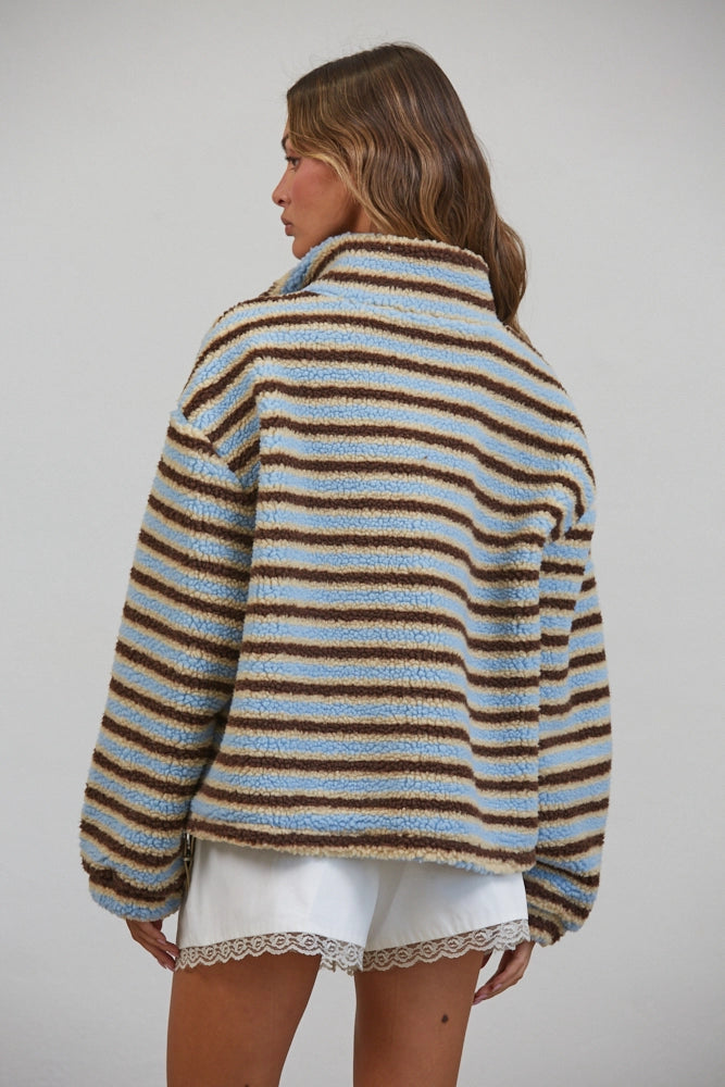 A woman shown from the back wearing a fuzzy striped cardigan with light blue, beige, and brown horizontal stripes, paired with white shorts, against a plain light gray studio background.