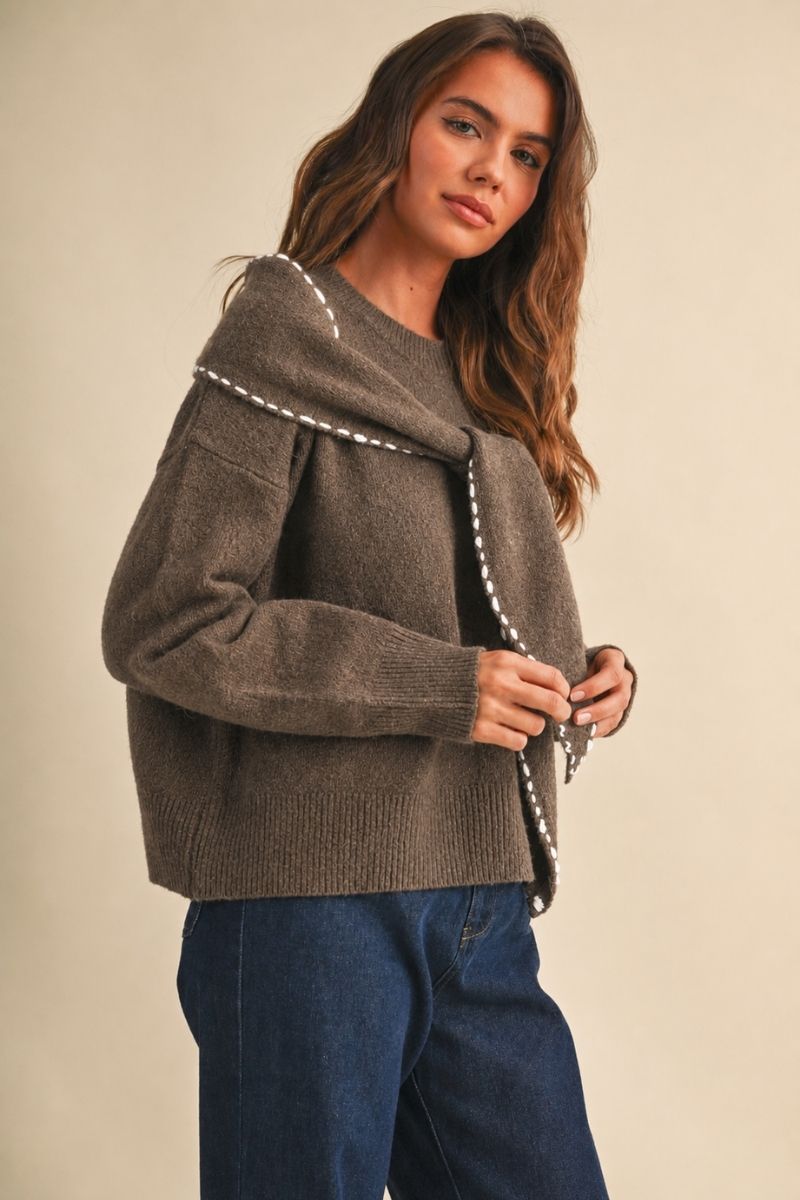 A woman wearing a brown knit sweater with a wide draped collar and white beaded trim, paired with blue jeans, against a plain beige background.