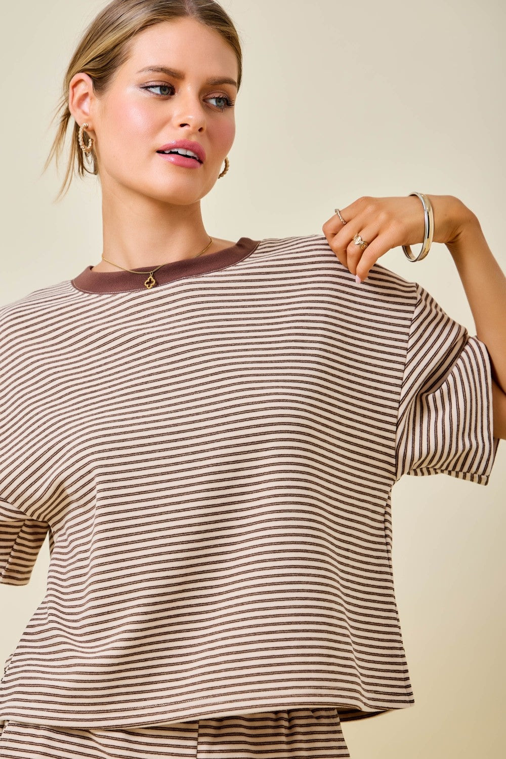 A woman wearing a brown and beige horizontally striped short-sleeve top with a solid brown crew neck, posed against a solid light beige background.