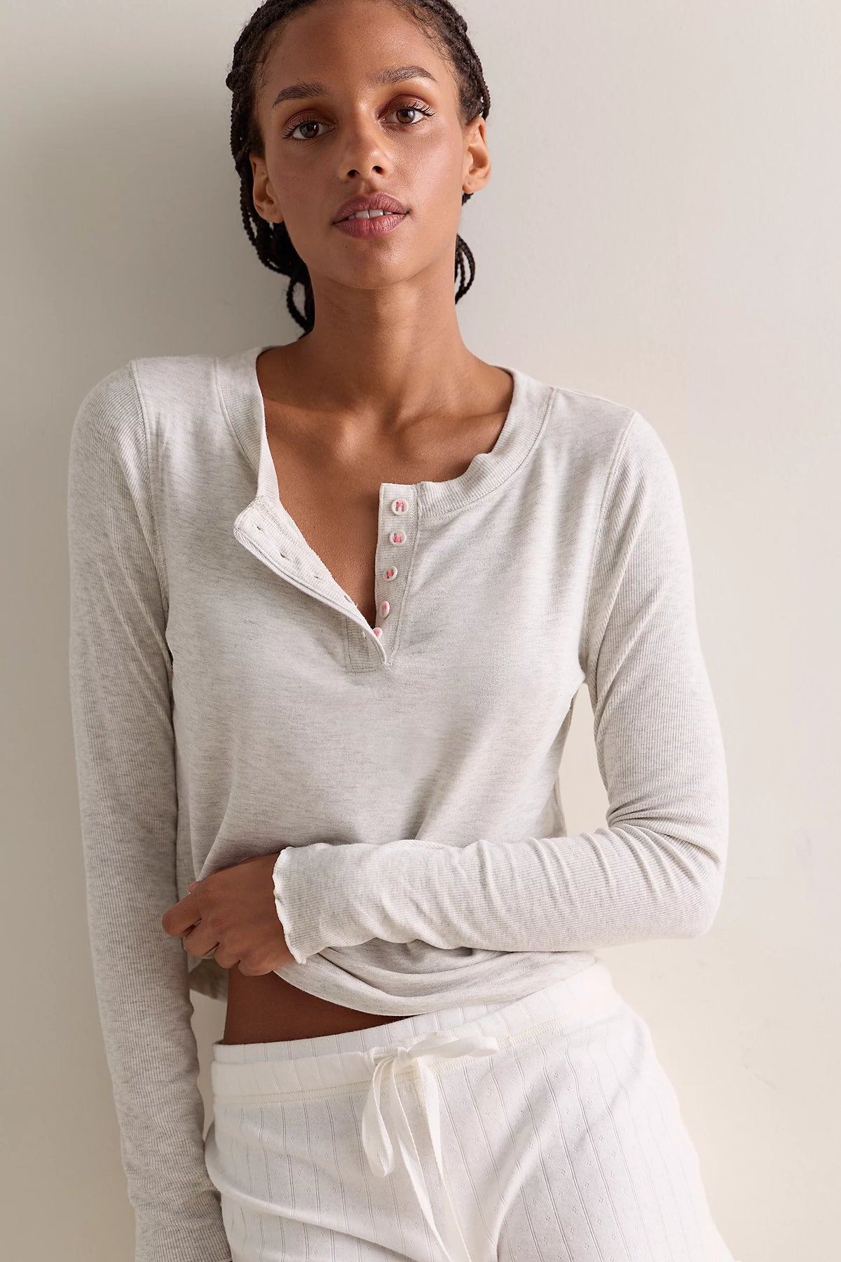 A woman wearing a light heather gray long-sleeve henley top with a buttoned split neckline and white drawstring pants, posed against a plain light beige wall.