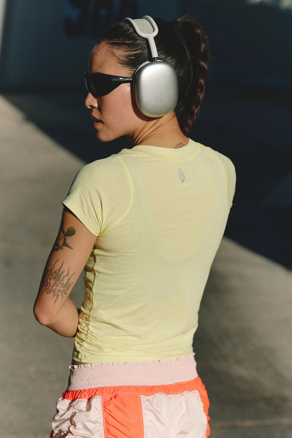 A woman seen from behind wearing a pale yellow athletic t-shirt, orange and white shorts, black sunglasses, and silver over-ear headphones in an outdoor setting.