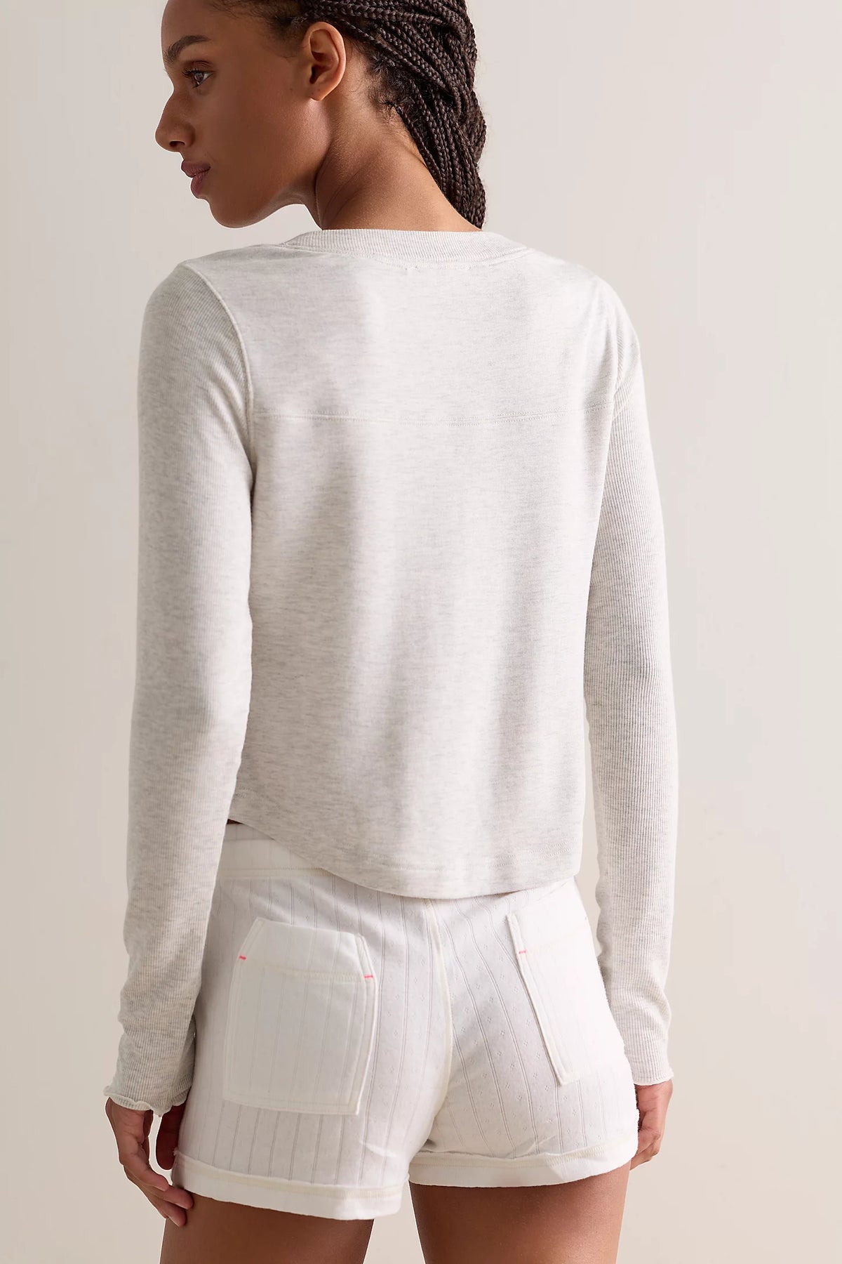 A woman shown from the back wearing a light heather gray long-sleeve cropped top and white shorts against a plain light beige studio background.