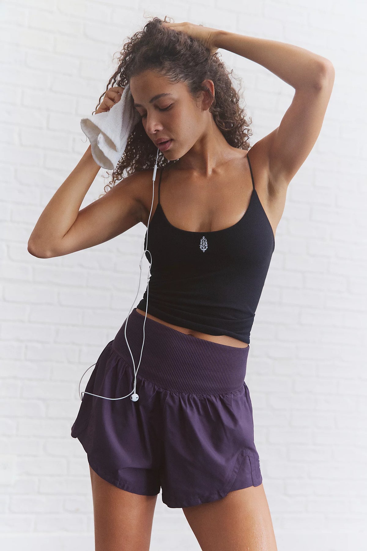 A woman in workout attire posing against a white brick wall, wearing a black tank top and purple athletic shorts, with white earphones and a white towel visible.