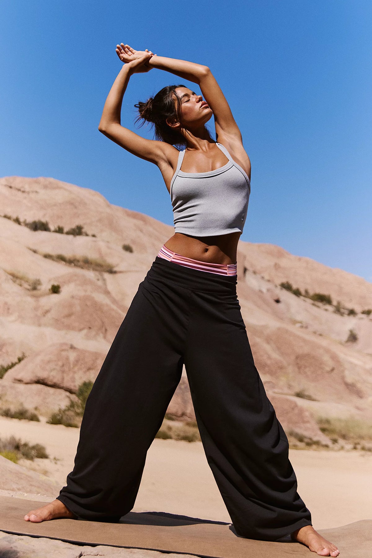 A woman in a desert landscape wearing a light gray ribbed tank top and black wide-leg yoga pants with a pink waistband, stretching on a yoga mat under a clear blue sky.