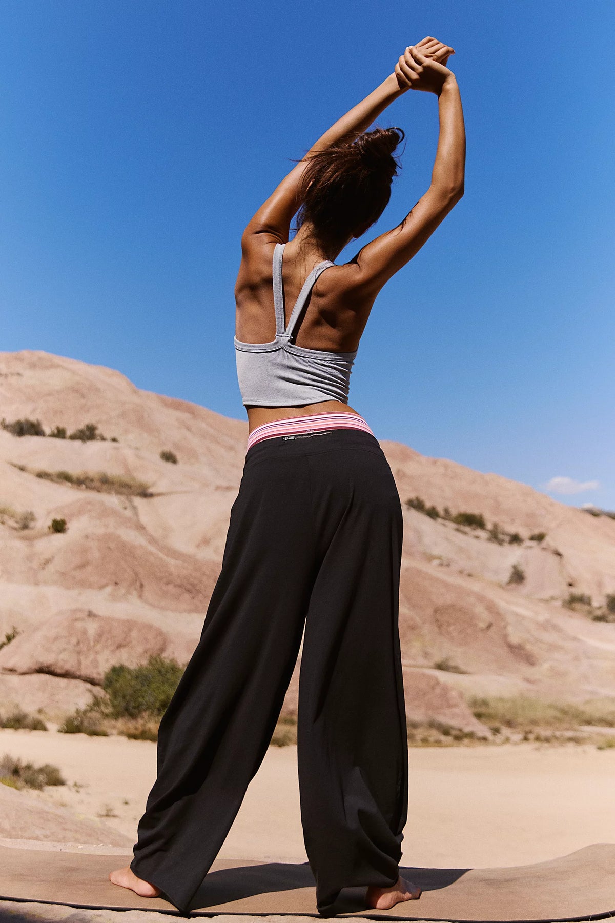 A woman in a gray sports bra and black wide-leg yoga pants poses from behind on a desert trail under a clear blue sky.