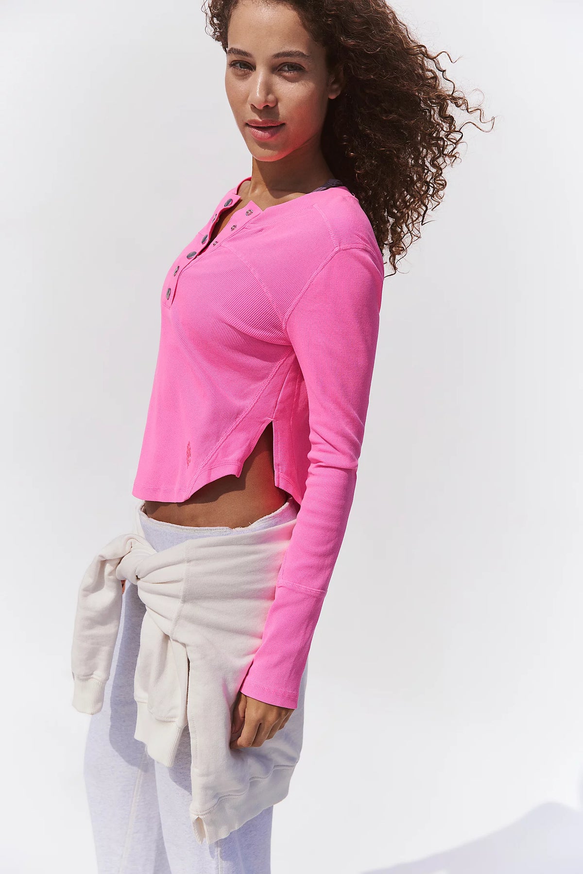 A woman wearing a bright pink long-sleeve cropped henley top with side slits, paired with white pants and a white sweatshirt tied around her waist, against a plain white background.