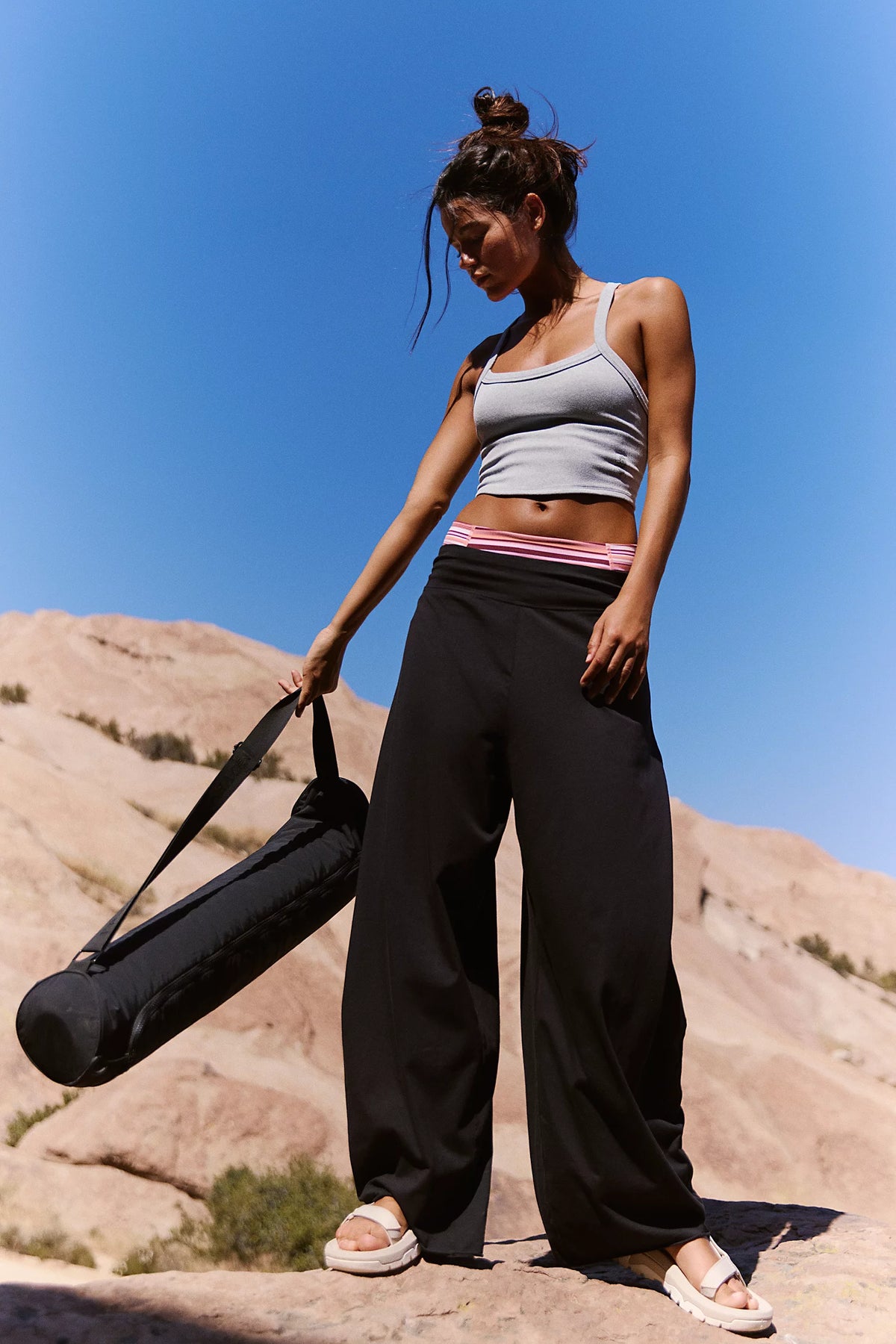 A woman stands outdoors on a rocky desert landscape under a clear blue sky, holding a black yoga mat bag. She is wearing a light gray ribbed tank top, loose black yoga pants with a pink and white striped waistband, and white platform sandals.
