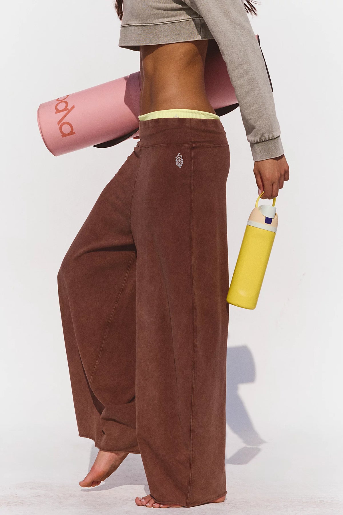 Person wearing brown wide-leg pants holding a pink yoga mat and yellow water bottle on a white background