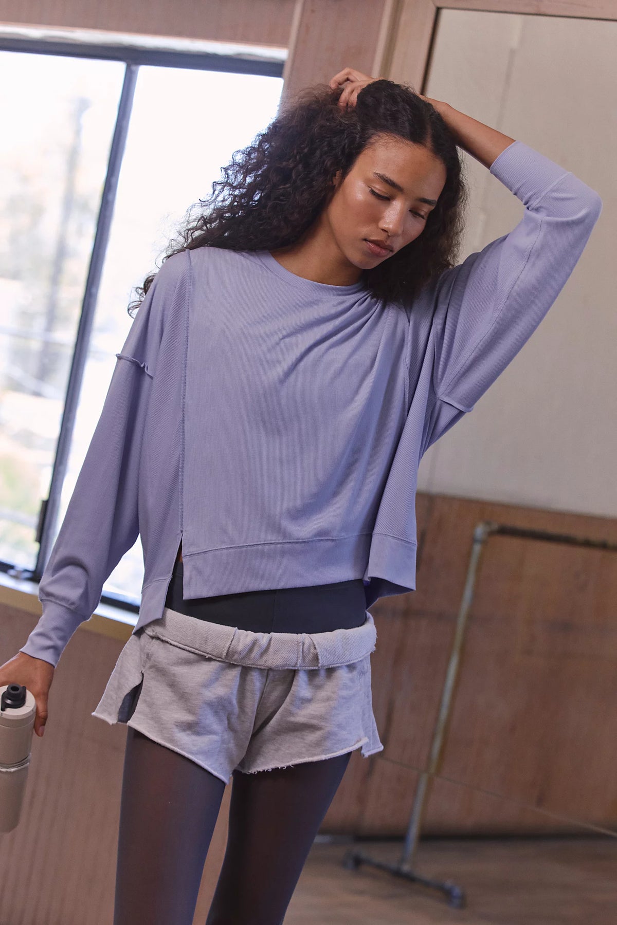 A woman in a studio setting wearing a lavender long-sleeve cropped sweatshirt, light gray shorts, and black tights, holding a white bottle.