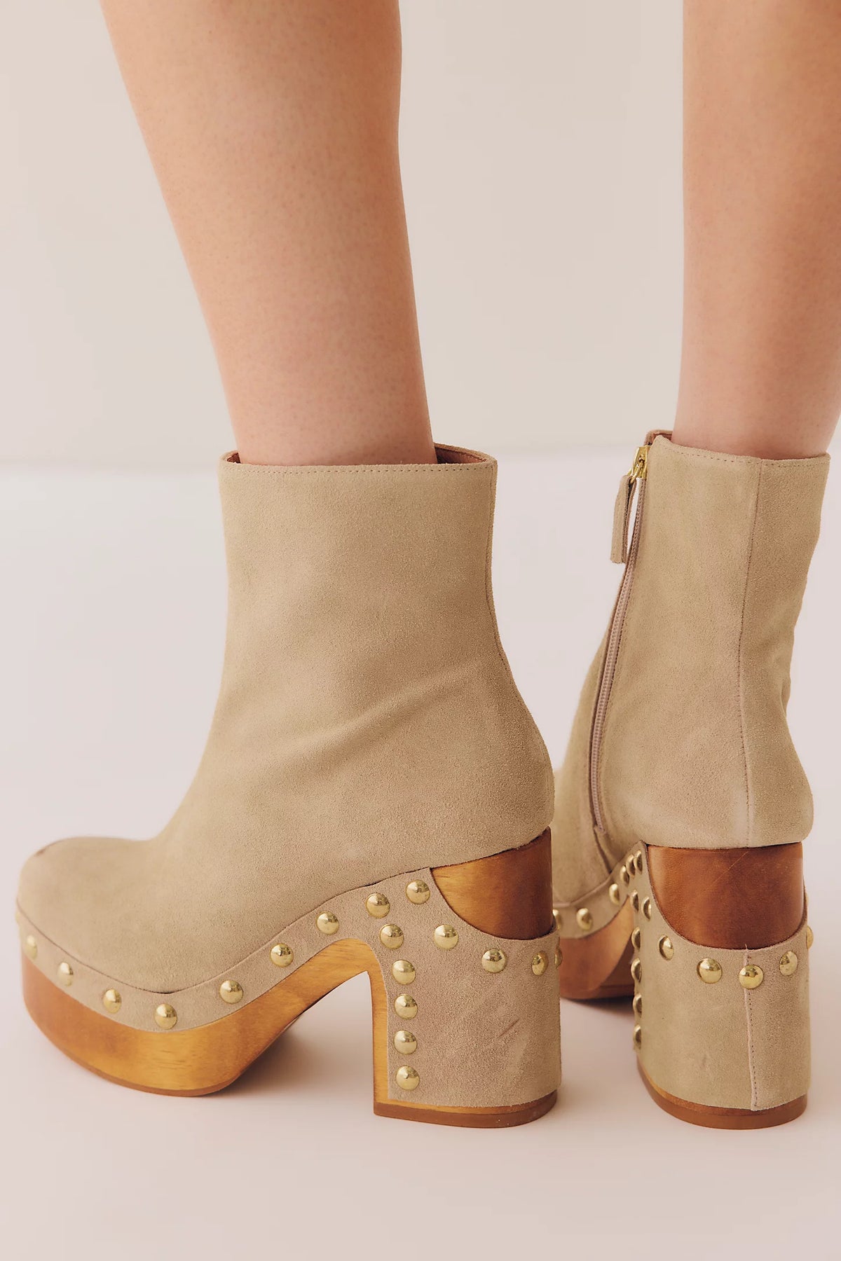 Beige suede ankle boots with chunky platform soles, gold-tone studs, and stacked wooden heels, shown on a model against a light beige background.