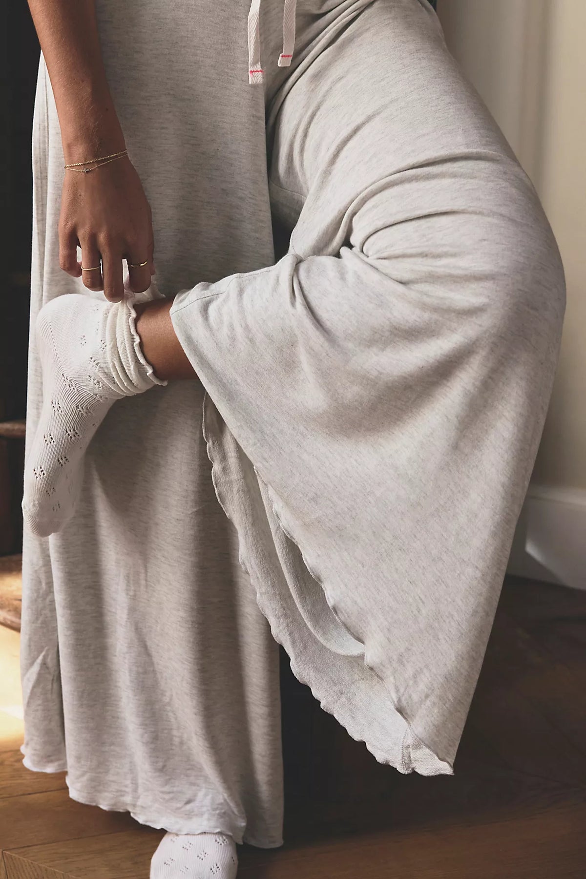 A person wearing a light heather gray lounge set, featuring a long-sleeve top and matching wide-leg pants, with white eyelet socks. The person is sitting on a wooden floor in a dimly lit indoor setting.