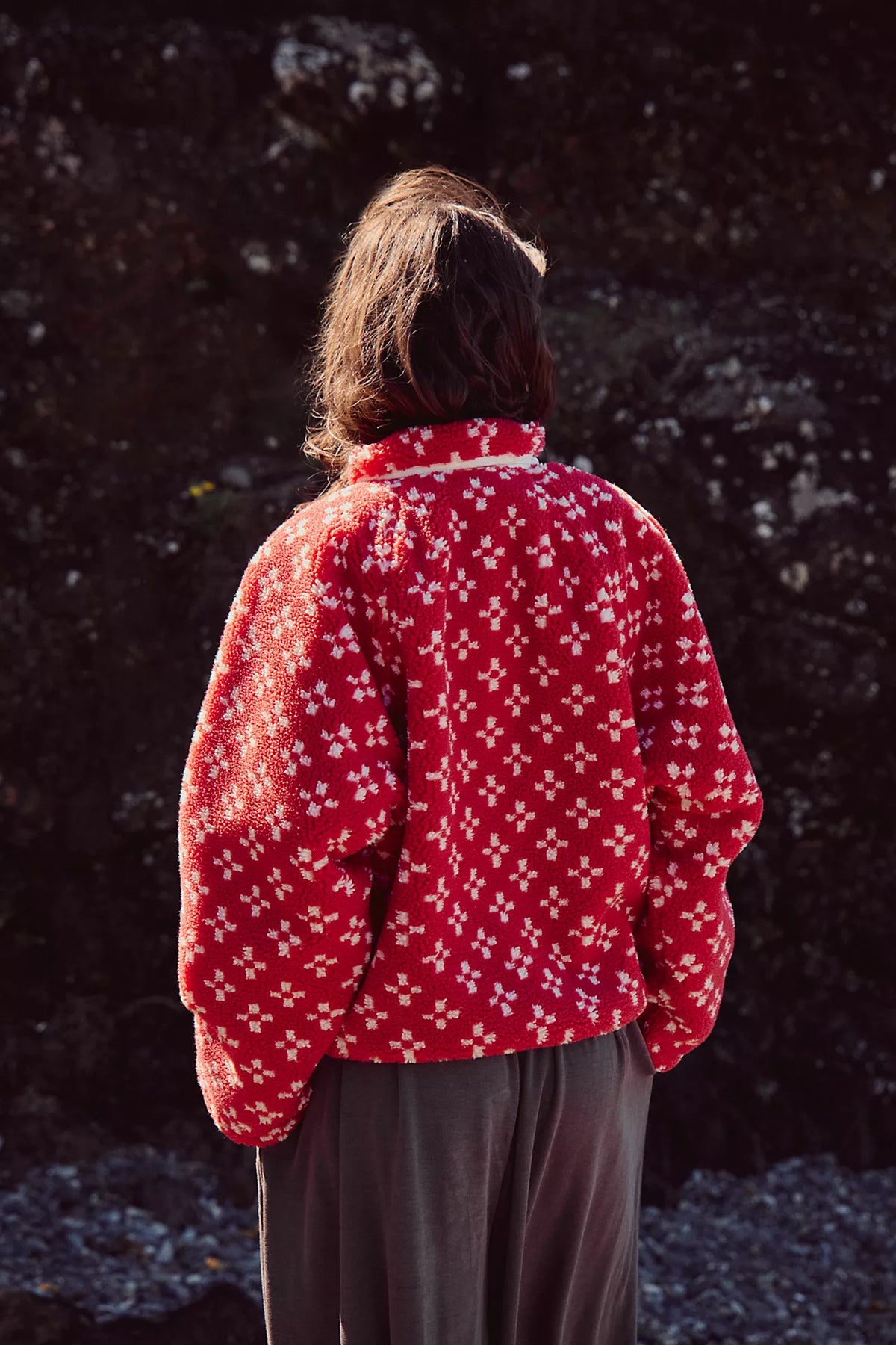 A person viewed from behind wearing a red fleece jacket with a white floral pattern and dark gray pants, standing outdoors against a dark rocky background.