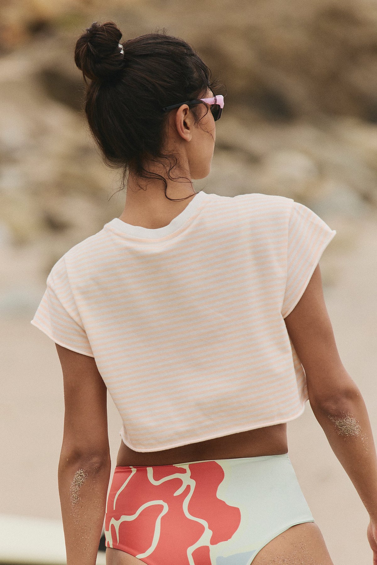 Woman in a beige crop top and colorful shorts standing on a beach.