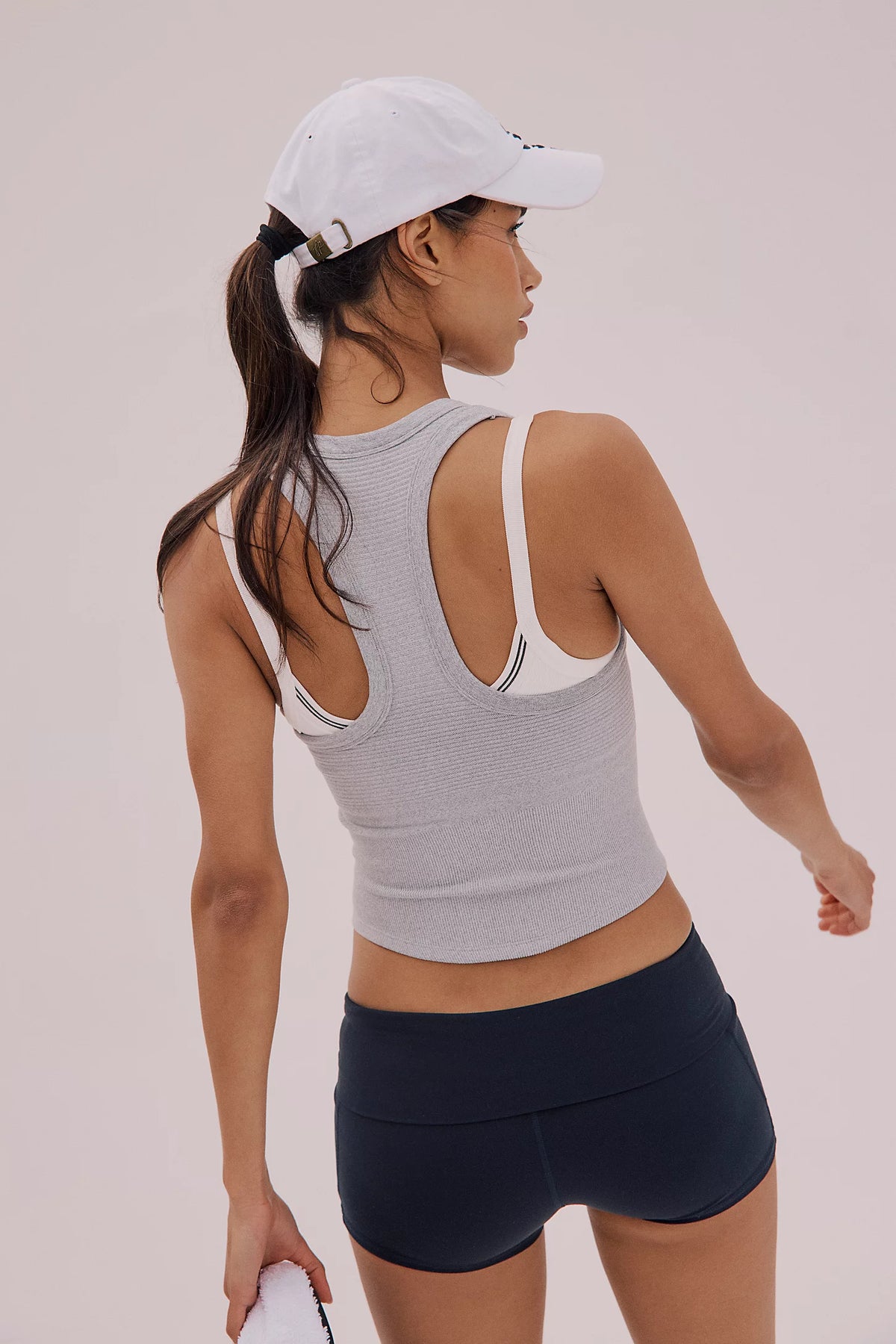 Woman wearing a gray sports bra and black shorts with a white cap on a light pink background