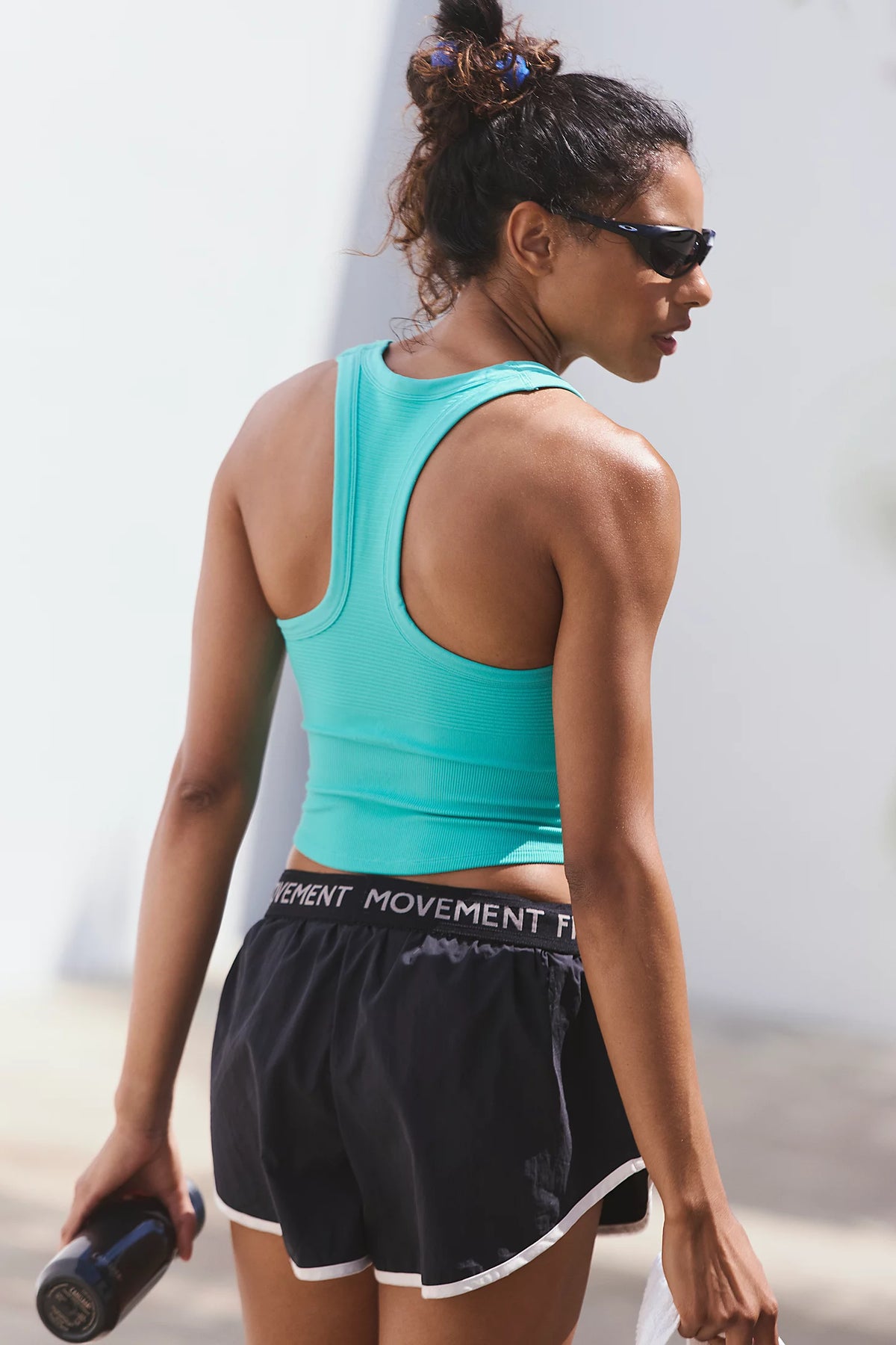 A woman wearing a turquoise athletic tank top and black shorts with white trim, shown from the back outdoors. The shorts have a visible waistband that reads 'MOVEMENT'.
