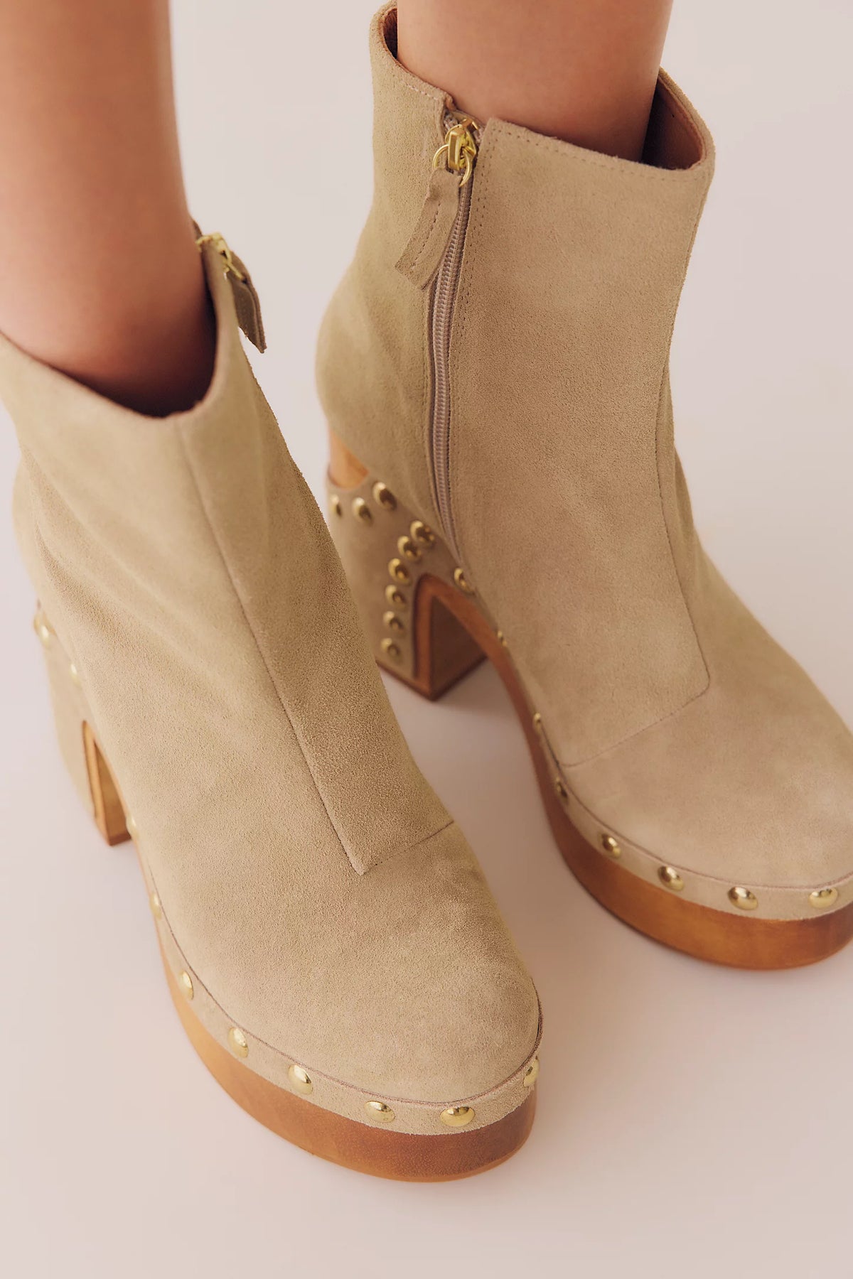 Beige suede ankle boots with gold-tone studs and a block heel, shown on a model's feet against a light beige background.