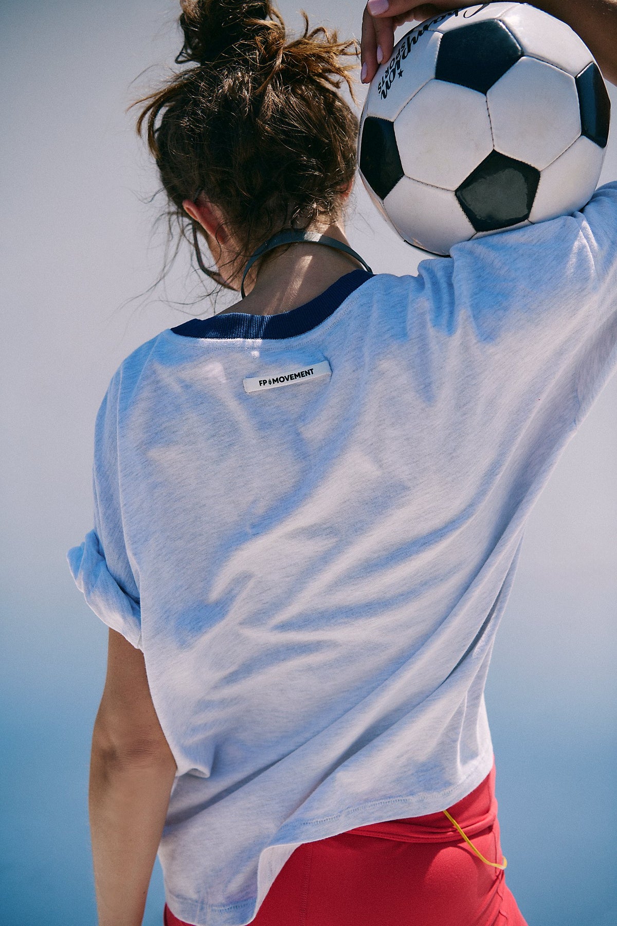Person wearing a light blue shirt with a visible brand logo, holding a soccer ball against a clear sky.