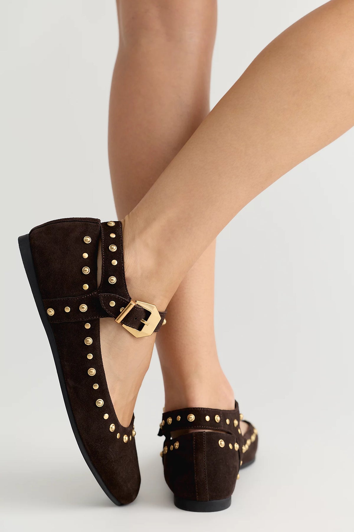 A pair of dark brown suede Mary Jane flats with gold-tone studs and buckles, shown on a model's feet against a light gray background.