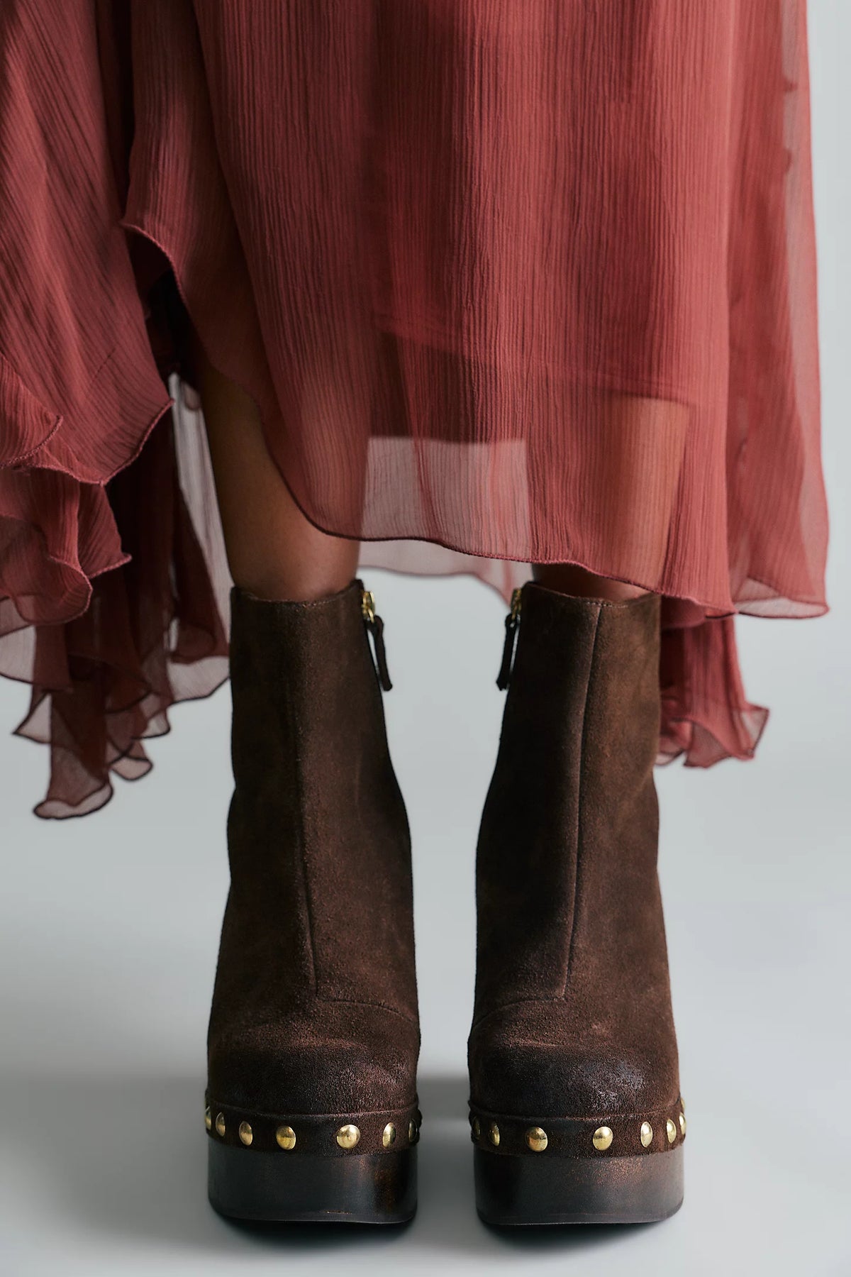 A pair of dark brown suede ankle boots with gold-tone studs on the platform soles, worn with a dusty rose chiffon skirt against a light gray background.